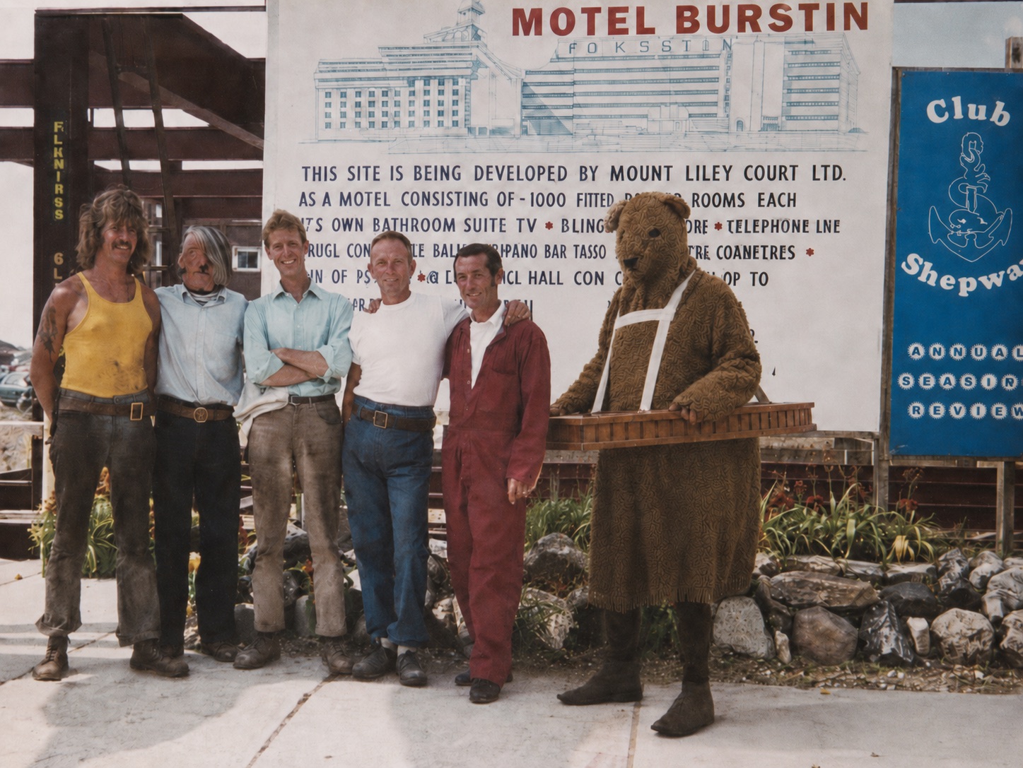 The Bear standing beside workers near a Hotel Burstin site board